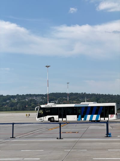 An airport shuttle bus, identified by the text "cobus 3000" and the number "2066", is parked on the tarmac. The bus is white with blue and white striped accents along its side, and a red logo with a white "LS" is visible on the front. A person in a high-visibility vest can be seen through the driver's side window. The bus is positioned on a paved surface with white line markings, typical of an airport runway or taxiway.
In the background, a line of trees creates a dense forest, with houses nestled among them, suggesting a location near a populated area adjacent to the airport. A tall pole with what appears to be a windsock or a wind direction indicator is present in the mid-ground, to the left of the bus. The sky is predominantly blue with scattered white clouds, indicating fair weather. The scene is illuminated by natural light, suggesting it is daytime. There are no overt signs of a specific event or activity taking place; the bus appears to be stationary. The location is identified as Balice, Poland.