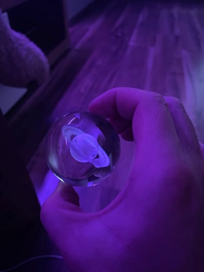 A hand holds a clear glass orb containing a miniature representation of the planet Saturn. The orb is illuminated by a purple light source, casting a vibrant glow on the surrounding environment. The background consists of a wooden floor and a dark, indistinct shape. The image appears to be taken indoors, possibly in a dimly lit room, with the purple light creating a dramatic and otherworldly atmosphere. There is no discernible location in Gliwice, Poland.