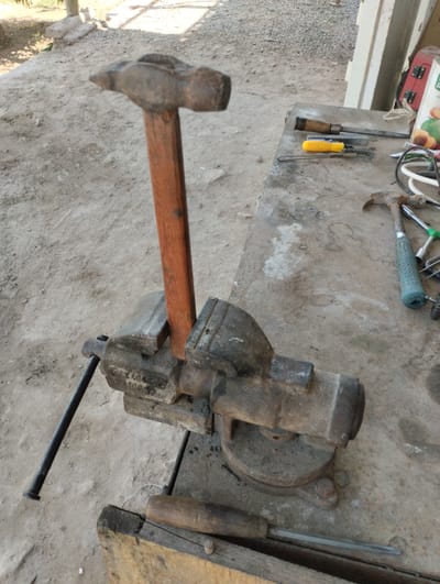 A close-up, high-angle shot shows a workbench in Yangibazar, Uzbekistan, cluttered with tools. In the foreground, a large, rusty vise grips the wooden handle of a heavy hammer, its metal head angled upwards and to the left. To the right, a concrete surface is covered with various items including a small hammer with a green textured handle, a metal file, a wire brush, a yellow-handled tool, and what appears to be a small red electrical box with visible wiring. The ground surrounding the workbench is dirt and gravel. The lighting suggests it is daytime.