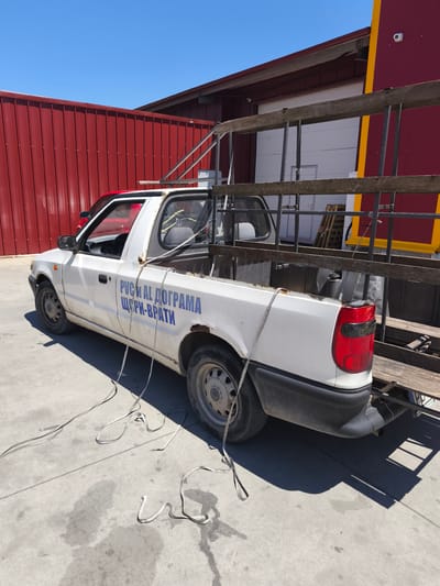 A white pickup truck is parked on a concrete surface outdoors. The truck has a red cab roof and blue lettering on its side that reads "РУСИ И АЛ ДОГРАМА ЩОРПИ-ВРАТИ". A metal ladder is loaded in the truck bed and secured with straps that also lie on the ground. The truck is parked in front of a red corrugated metal building with a yellow stripe and a portion of a dark red building with a white garage door visible in the background. The sky is clear and blue. The scene appears to be in an industrial or commercial area.