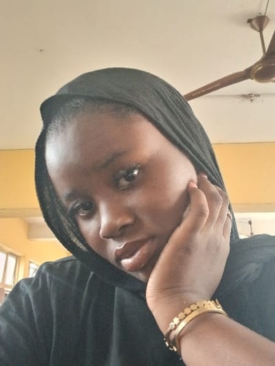 This image captures a young individual, possibly female, of African descent, posed in a three-quarter frontal view looking towards the camera. The individual wears a dark, possibly black, head covering and a dark-colored top. The right hand is raised, with fingers partially curled, resting against the right side of the face/jawline. Multiple gold-toned bracelets adorn the right wrist, including a wide cuff with a decorative pattern and a thinner bangle. The lips exhibit a glossy appearance.
The scene is indoors, located in the city of Awjaw, Nigeria. The background features a light-colored ceiling, from which a brown, three-bladed ceiling fan is mounted in the upper right quadrant. A wall section to the left appears to be painted a pale yellow or light orange. A window structure is partially visible on the far left in the background, indicating a source of external light. The overall lighting is consistent with an interior setting.