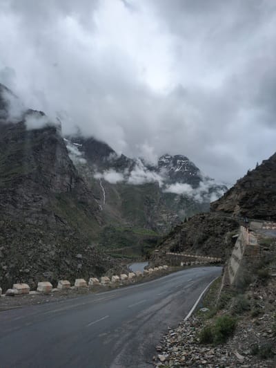 The image captures a winding mountain road in Gemur, India, under a cloudy sky. The road, paved with asphalt, curves from the foreground to the mid-ground, flanked by rocky terrain and a concrete barrier on the right. The barrier has an aged, textured appearance, with intermittent white and orange markers. Small rocks and sparse vegetation cover the slopes on either side of the road. In the background, imposing, rugged mountains rise, partially obscured by thick, gray clouds. A thin waterfall cascades down one of the mountain faces, adding a dynamic element to the otherwise static landscape. The overall atmosphere is somber and dramatic due to the overcast weather. There are no discernible people or vehicles in the immediate foreground, though a distant hint of cars can be observed on the road in the mid-ground.