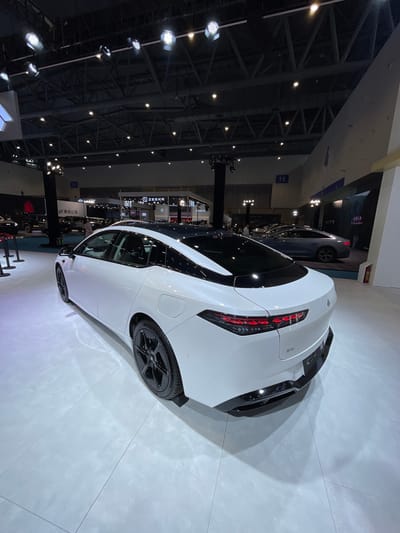 The image shows a white car, a Zeekr 001, parked on a white tiled floor in a large, brightly lit indoor space. The car's rear right side is in view. The car's front is partially visible, and there is a second, black car in the background to the left of the white car. There is a third car visible on the left side of the image, but it is obscured. The car is located in a large exhibition hall, with a ceiling constructed of metal beams. The hall is empty except for the cars and a few people walking around. There are no other objects or features in the image.  The car is located in the Hangzhou International Expo Center at coordinates 30.272 latitude and 120.416 longitude.