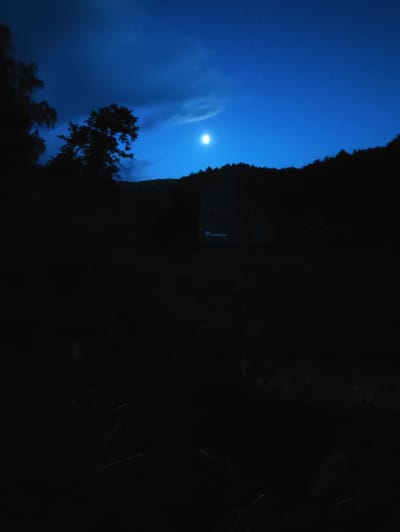 A dark, nighttime landscape with a prominent full moon shining in a deep blue sky. The moon is slightly off-center to the left, partially obscured by a wispy cloud. Silhouetted trees and a rolling hill form the horizon line. In the foreground, a dark, indistinct area suggests a campsite with a tent visible as a faint outline against the hillside. The tent appears to have a logo with visible text, though it is too blurry to read clearly. The overall mood is serene and tranquil, with the moonlight casting a cool, ethereal glow on the scene. The time of day is clearly night, and the weather appears clear aside from the light cloud cover around the moon. The location context suggests a rural or natural setting in Kul'metovo, Russia.