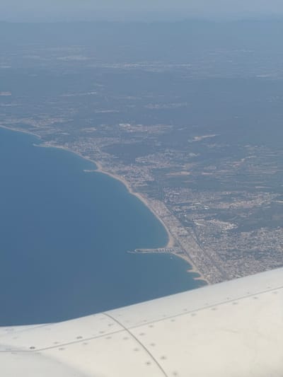 This media file captures an aerial view of a coastal city and the sea, likely taken from an airplane window. The scene shows a dense urban area extending along a coastline, meeting a calm, blue body of water. The city appears to have a mix of buildings, with some areas showing more concentrated development. A prominent feature is a long, sandy beach curving along the shore, with evidence of man-made structures like piers or breakwaters extending into the water. A road or highway runs parallel to the coastline, separating the urban sprawl from the beach.
The perspective is from a high vantage point, looking down at an angle. Part of an airplane's wing or fuselage is visible on the right side of the frame, characterized by its white, metallic surface with visible rivets and panel lines. The lighting suggests it is daytime, likely with clear or partly cloudy weather, as the overall scene is well-illuminated and the colors are vibrant. The image is oriented such that the coastline runs diagonally across the frame.
The location context provided places this scene in Vilanova i la Geltrú, Spain. This information suggests the coastline depicted is part of the Mediterranean coast of Catalonia. There is no visible text in the image. The overall mood conveyed is one of travel and observation, offering a sweeping panorama of a developed coastal landscape from above.