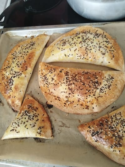 A close-up shot shows several baked pastries on a baking sheet lined with parchment paper. The pastries are golden brown and dusted with a mixture of sesame seeds, flaxseeds, and possibly other small seeds. They appear to be folded or crescent-shaped, with some cut into triangular pieces. The background is blurred, showing a stovetop and a metallic pot, suggesting a kitchen environment. The image does not contain any elements that would allow for its localization in Andzhero-Sudzhensk, Russia, or any other specific city.