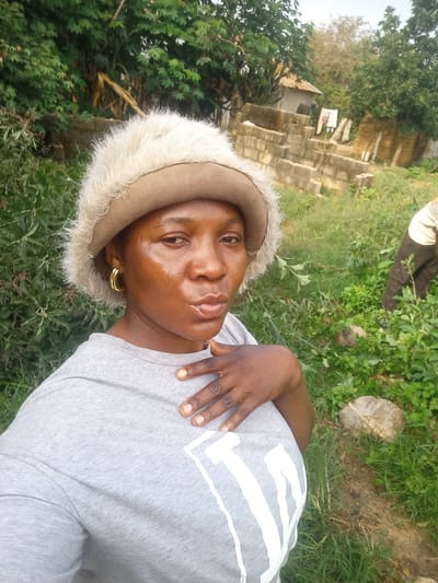 A woman wearing a furry tan bucket hat and a grey t-shirt with a white graphic is captured in a selfie outdoors. She is looking at the camera with her mouth slightly pursed and has her right hand on her chest. Her left ear is adorned with a gold hoop earring. Behind her, a person in the background appears to be tending to some vegetation. The setting includes green foliage, a structure built of concrete blocks, and a modest building with a corrugated roof. The environment suggests a rural or semi-urban area, possibly within the city of Mangu Halle, Nigeria.