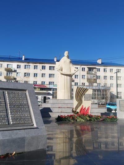 The image captures a monument in Zhatay, Russia. The monument consists of three distinct parts: a marble statue of a robed woman, a marble memorial slab with stylized red flame accents, and a gray granite plaque bearing text in Russian. Red flowers are laid at the base of the memorial. The surface in front of the monument is reflective and wet, mirroring the statue and the surrounding structures. In the background, a multi-story apartment building with white walls and a blue roof is visible under a clear blue sky. A blue car is parked on the street behind the memorial. The memorial plaque features names inscribed in Cyrillic script.