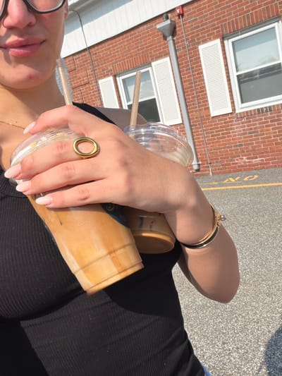 A woman stands outdoors, holding two iced coffees. She is wearing a black tank top and jewelry, including a gold ring on her right index finger and a gold bracelet on her left wrist. The background consists of a brick building with white-shuttered windows and a paved area with yellow markings. The lighting suggests it is daytime. The specific location within Ridgewood, United States of America, is not determinable from the image.
