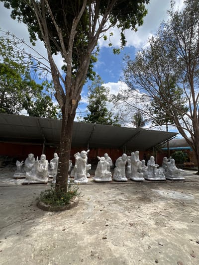 This image depicts an outdoor workshop or storage area in Đăk Mar, Vietnam, under a partly cloudy blue sky. The foreground features a large, multi-trunked deciduous tree with sparse foliage, surrounded by a circular concrete planter containing small yellow-flowering plants. The ground consists of uneven, light grey concrete pavement with scattered debris and shadows.
In the mid-ground, under a long, open-sided structure with a corrugated metal roof, a collection of approximately 15-20 white marble or stone statues are arranged in rows. These statues depict human figures in various poses, some seated, others standing or kneeling, consistent with religious or mythological representations. Many of the statues are partially covered in clear plastic sheeting or white fabric for protection. They rest directly on the paved ground, with some appearing to be on wooden pallets.
Beyond the covered structure, lush green trees and darker foliage are visible, along with glimpses of reddish-brown structures and potentially another building or fence in the far background. Another tree with a lighter trunk and fewer leaves stands on the right side of the frame. The scene is illuminated by bright daylight, casting distinct shadows.