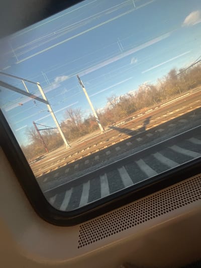 View from a train window showcases parallel tracks and overhead lines against a bright sky. Trees line the distant landscape, their bare branches hinting at autumn or winter. The window frame is black, and a panel with small circular perforations is visible on the right, suggesting the interior of the train. The tracks are marked with alternating dark and light stripes.