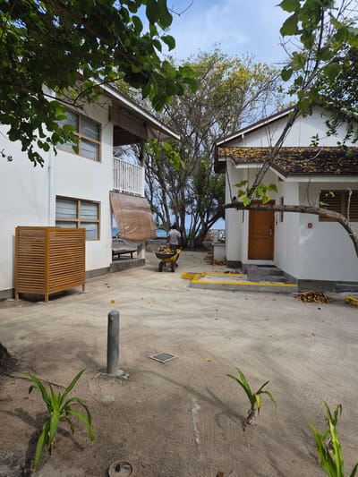 The image shows a partially obscured view of buildings and a walkway on Huraa, Maldives. A man is seen pushing a wheelbarrow near a building with a brown wooden door. The buildings are white with wood-framed windows. Trees are present, some obscuring the building. The walkway is made of concrete and sand. Some scattered yellow leaves are seen on the walkway and on the rooftops. There is also an outdoor area with an elevated platform covered with tarp.