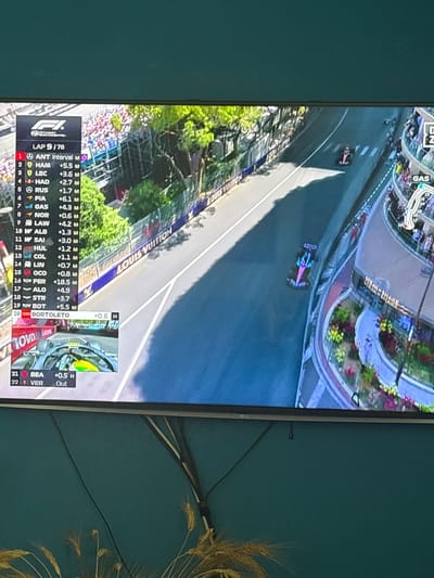 The image displays a flat-screen television mounted on a matte teal wall in a residence located in Urbanización Encinares De Alcolea, Spain. The television broadcasts a Formula 1 race, which appears to be the Monaco Grand Prix, indicated by the distinctive street circuit environment, multi-level spectator balconies, and "LOUIS VUITTON" branding on trackside barriers. The screen shows "LAP 9/78" and a leaderboard on the left displaying driver positions, with "ANT" in 1st, "HAM" in 2nd, and "VER" marked as "Out" in 22nd. Two race cars are visible on the track: one in the mid-ground with a pink, blue, and purple livery, and another in the background in black and pink. A smaller picture-in-picture window at the bottom left displays an onboard view from a car, showing a yellow and green helmet. A stylized track map with a "GAS" indicator is partially visible on the upper right. The television is an LG brand, identifiable by a logo on its lower bezel. Beneath the television, a decorative arrangement of dried wheat stalks is present. A single black cable extends vertically from behind the top center of the television.