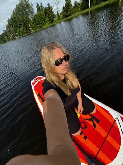 A blonde woman wearing black sunglasses and a black t-shirt and shorts is sitting on a red and orange striped paddleboard on a body of water. The paddleboard has black lettering that appears to spell out "KO". The water is dark and appears to be slightly rippled. In the background, there is a shoreline with green trees and a partially cloudy sky. The image is taken from a high angle, looking down at the woman and the paddleboard. It appears to be a sunny day. The location is Zamosh'e (Zaklinskoe S/P), Russia.