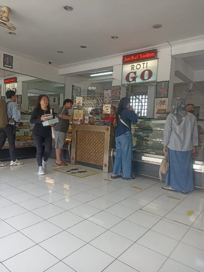 The image shows the interior of a bakery called "Jendral Sudirman Roti Go" located at the coordinates -7.428, 109.247 in the city of Purwokerto, Indonesia. The bakery has a tiled floor with yellow tape arrows directing customers. There are five people inside: two customers waiting at the counter, one customer near the counter to the left, one cashier, and one person standing further back to the left. There is a large display case with prepared goods in the center of the image, while the counter is on the left. The bakery has a white ceiling with recessed lighting and a sign with the name of the bakery in red lettering. The walls are white and there are several posters on the wall behind the counter. There is a large window to the right of the display case showing a glimpse of the street outside. A person in the back is wearing a mask. There are some bags on the counter near the display case. The bakery is well-lit.