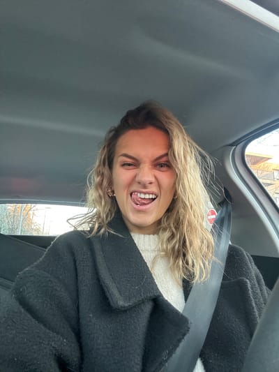 Inside a vehicle, a woman with blonde, wavy hair and a black coat is captured at an angle. She wears a white sweater, a black seatbelt, and a light-colored pair of earrings. Her facial expression is exaggerated, one eye squinted, mouth open, and tongue partially extended. The interior of the car includes a gray ceiling and glimpses of a window with buildings visible outside.