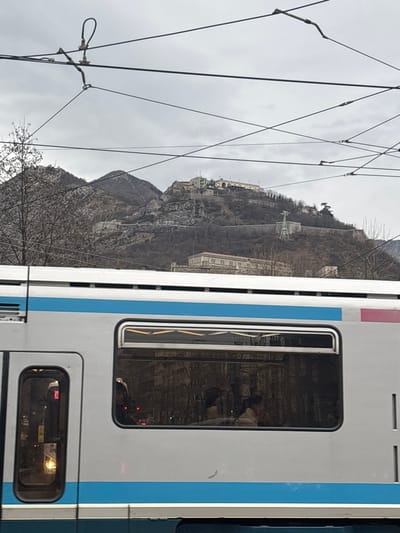 The image, taken in Grenoble, France, captures a side view of a white train or tram with a horizontal blue stripe, occupying the right side of the frame. Its windows are dark and reflective; a larger window on the upper right reflects indistinct interior shapes and possibly the outline of a person. A smaller window below it shows a yellow sign with the number "10".

In the background and to the left, a steep, rocky mountain rises under an overcast, light grey sky. The mountain is covered with numerous bare trees, indicating a winter or early spring season. Perched high on the mountain's slopes are several buildings and structures, strongly resembling the historic Bastille fortress overlooking Grenoble. A notable green structure, possibly part of a monument or an installation related to the cable car system, is also visible on the mountainside.

Numerous black overhead power lines crisscross the upper left portion of the image, appearing diagonally against the cloudy sky and partly in front of the mountain. The overall scene depicts a typical urban-mountain interface found in Grenoble, with its distinctive transportation network and famous landmark. No specific activity or event is clearly depicted beyond the presence of the train/tram and the landscape.
