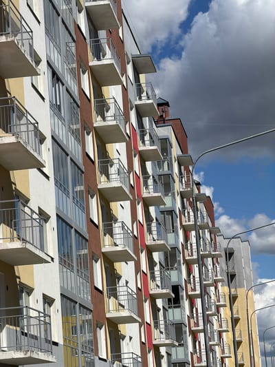 The image shows the facade of a multi-story building in Янино-2, Russia. The building has a stepped design, with each level slightly recessed compared to the one below. Each level features a row of identical rectangular balconies with minimalist metal railings and concrete dividers. The building's exterior is painted in horizontal stripes of white, yellow, brown, and red. Small windows are also visible between the balconies. The sky is partly cloudy, with blue patches visible behind the building. A metal cable runs across the upper part of the image.