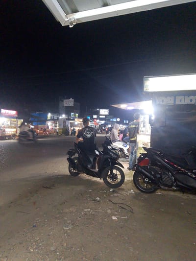 This is a nighttime, outdoor shot of a street scene in Gampongpaya, Indonesia. The scene is illuminated by streetlights and lights from various shops and food stalls. In the foreground, several motorcycles are parked on a dirt and gravel road. One motorcycle is prominent in the lower right, showing its rear wheel and exhaust. Further back, a man is seated on a black motorcycle, facing away from the camera, wearing a black t-shirt with a circular logo.
To the left of the road, several food stalls are visible, with bright neon signs. One stall has "DIMSUM Premium" written on it, with "2.000" visible below, likely indicating a price in Indonesian Rupiah. A blurry image of a person on a motorcycle passes in front of these stalls. In the background, there are more buildings and lights, suggesting a bustling street or market area.
To the right, a dark building with a brightly lit awning stands out. A sign above the awning displays white text that is partially obscured but appears to read "YOGI KUTO" or something similar. Beneath the awning, two men are interacting, one is standing and looking at merchandise displayed on a shelf. A woman wearing a hijab is seated on a motorcycle near them. The overall atmosphere appears to be lively and active, typical of a night market or street food area. The time of day is clearly night, and the ground suggests it might be a bit dusty or unpaved.