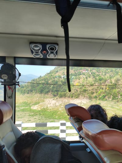 A POV shot from inside a bus shows passengers sleeping as the bus travels through a mountainous landscape. In the foreground, the backs of three passengers are visible, with two appearing to be asleep, their heads resting on their shoulders or the seats. The bus interior features orange and cream colored seats, a ceiling with speakers, and a small fan mounted to the left. Through the large windows, a steep, tree-covered hillside is visible, dotted with small houses. The landscape is illuminated by bright daylight, suggesting it is daytime. The scenery is consistent with the mountainous terrain found in and around Bilaspur, India, a city known for its natural beauty and surrounding hills.