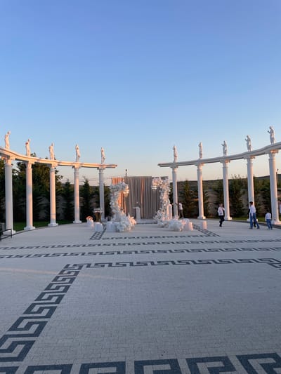 The image depicts an outdoor event space with neoclassical architectural elements, located in Ratuș, Moldova. The scene is characterized by a colonnade with statues of figures standing atop white pillars. In the foreground, a paved courtyard features a geometric pattern in grey and black stone. To the right of the courtyard, two white balloon arches are erected, one flanking a draped partition and the other standing independently. A small round table with glasses is set near the arches. Several people are present in the scene. Towards the right side of the image, two adults and two children, all dressed in white, are walking. Further to the left, another adult is seen near the draped partition, seemingly interacting with the setup. The environment is bathed in the warm light of a sunset, casting long shadows across the pavement. Trees form a backdrop behind the colonnade, and a gently sloping hill is visible in the distance under a clear blue sky.
