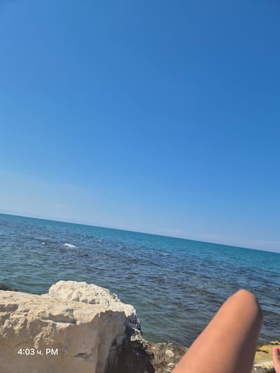 An outdoor, low-angle, medium shot captures a bright, sunny day at the sea. A person's bare leg and knee are visible in the bottom right corner, with a glimpse of a red toenail. In the foreground, large, light-colored rocks and smaller dark rocks are situated on the shoreline. The sea is calm with gentle ripples, transitioning from clear, shallow water near the shore to a deeper blue further out. A faint, small cloud is visible in the distance, and a hazy line of land or mountains can be seen on the horizon under a clear blue sky. The image contains a timestamp in the bottom left corner reading "4:03 Ч. PM." The scene appears to be a relaxed setting by the sea, possibly a beach or rocky coastline.