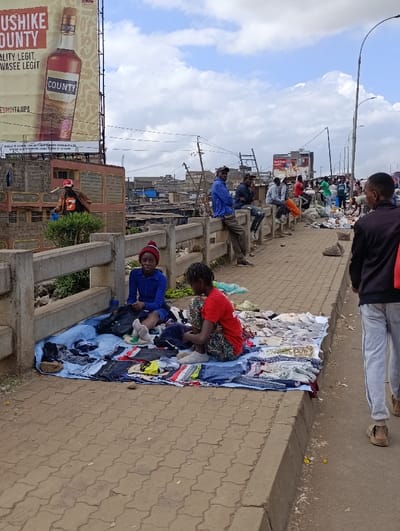 This image captures a lively street scene in Kiuu ward, Kenya, featuring informal commerce under a partially cloudy sky during daytime.
In the foreground, two young girls are seated on a blue tarp laid out on the patterned pavement, displaying a variety of clothing items like t-shirts, jeans, and socks for sale. The girl on the left wears a blue top and a red beanie, while the girl on the right is in a red t-shirt and patterned pants, both appearing to be engaged with their merchandise. To the right, a man in light-colored sweatpants and sandals walks past, his back to the viewer.
The midground features a concrete barrier, likely part of a bridge or overpass. Several men are sitting or leaning on this barrier, observing the activity on the busy street. Further down the street, more people and piles of goods or discarded items suggest a bustling market environment.
The background reveals a dense collection of buildings and structures, typical of an urban or peri-urban settlement. Utility poles and overhead lines are visible against the sky. A prominent billboard in the upper left advertises an alcoholic drink called "COUNTY." Visible text on the billboard includes "USH*KE COUNTY," "QUALITY LEGIT, WASEE LEGIT," and "COUNTY" printed on the bottle. Additional smaller text, including "ESHOTAJPA" and a disclaimer, is also present but less legible.
The scene conveys a sense of daily life and informal economic activity in a populated area. The clear daylight indicates it is likely late morning or early afternoon.