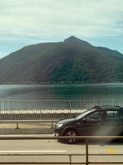 The image captures a scenic view from a bridge in Melide, Switzerland, overlooking Lake Lugano and a lush, tree-covered mountain. A black car is driven across the bridge, its occupants partially visible through the tinted windows. The lake's surface is calm, reflecting the soft light of the sky. A metallic railing lines the side of the bridge, separating the road from the water. The mountain dominates the background, its slopes densely forested, with a distinct peak at its summit. The sky above is a pale blue with scattered clouds.