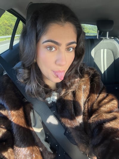 The image displays a female subject occupying the front passenger seat of a vehicle in Caminha, Portugal. She has dark, wavy hair, distinct dark eyebrows, and light brown eyes, looking directly at the camera. Her mouth is open, and her tongue is extended outwards. She is wearing a dark brown fur coat over a dark garment with a white lace collar and small, light-colored embellishments. A black seatbelt is securely fastened diagonally across her chest and shoulder. Direct sunlight illuminates the interior, creating sharp shadows on her clothing and the car's interior surfaces. Visible behind her is a dark gray passenger seat with lighter stripes on its backrest. Through the vehicle's windows, a road with a guardrail and undulating green, tree-covered terrain are discernible under a bright, clear sky.