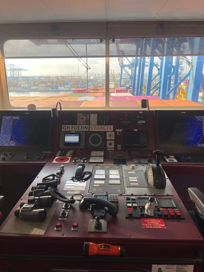 This media file depicts the interior of a ship's bridge, offering a view of the command console and a large window overlooking a busy port. The command console is filled with various instruments, screens, and controls, including radar displays, navigation equipment, and communication devices. A prominent label reads "GH FOEHN V7A4674". A red warning sticker states "DURING BOW THRUSTER OPERATION FLAP FOR VENTILATION TO BE OPENED."

Through the window, a vast container port is visible, characterized by towering cranes, stacks of colorful shipping containers, and other vessels. The text "ABIDJAIN TEA" is visible on the ship's exterior, suggesting the location of Abidjan, Ivory Coast. The sky appears overcast, hinting at potentially humid or rainy weather, common in tropical port cities. The overall scene suggests a working environment on a cargo ship, likely in port or preparing for departure. No people are clearly visible, but the presence of the advanced console implies the imminent activity of the ship's crew. The time of day is indeterminate, but the lighting suggests it could be daytime, with the overcast sky softening the light.