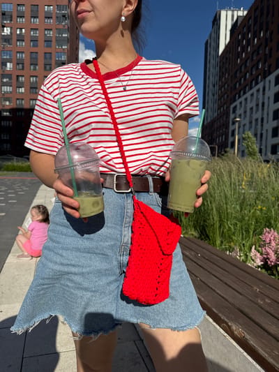 In an urban outdoor setting, a person is holding two clear plastic cups containing green iced beverages with green straws. They are wearing a red and white horizontally striped t-shirt, a denim skirt, and a red crocheted bag is slung across their body. In the background, modern buildings and a wooden bench are visible, along with lush greenery and flowers. Further in the distance, a young child in a pink shirt and shorts is seated on the ground. The scene appears to be located in Patrusheva, Russia, possibly in a residential area or a park with new construction.
