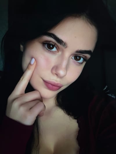The image is a close-up, indoor photograph of a young woman. She is positioned in the foreground, with her head slightly tilted and her left index finger touching her cheekbone, near her eye. Her eyes are brown and her eyebrows are dark and well-defined. Her lips are slightly parted, and she is wearing light pink lipstick. Her skin appears pale with a slight rosy blush on her cheeks. She has dark hair that frames her face. She is wearing a dark red top. The background is dark and indistinct, suggesting an indoor setting with low lighting. There are no visible landmarks or elements that would indicate a specific location in Belyye Berega, Russia, or any other city. The focus is solely on the woman's face and upper body.