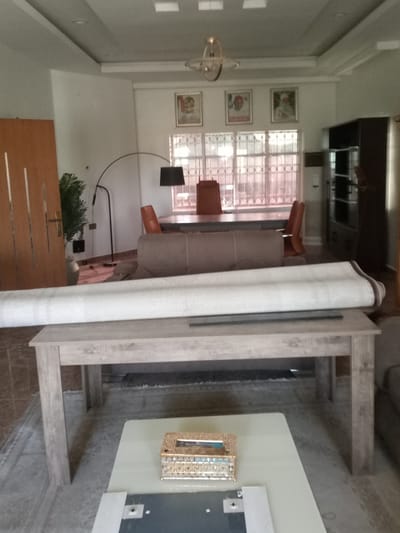 The image shows an interior scene, likely a living room or dining area in a residence in Hotoro North, Nigeria. In the foreground, a large rolled-up rug is placed on a light-colored wooden table. Behind the table, a sofa is visible, and further back, a dining table with chairs is set up. Several framed pictures hang on the wall above the dining area, and a tall floor lamp is positioned nearby. A closed wooden door is on the left side of the image, and a dark cabinet is present on the right. The overall lighting suggests it is daytime.
