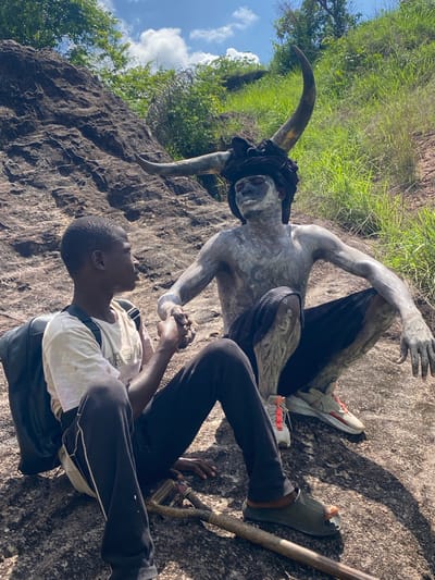 A man lies on his back on a rock surface in Ufueseni, Nigeria, with a horned figure lying nearby. The man wears a light-colored t-shirt, dark pants, and a black backpack. He holds the figure's hand. The figure, possibly a statue or a person covered in paint, has two large horns and dark clothing. The ground is rocky and surrounded by green vegetation. The sky is visible in the background with white clouds.