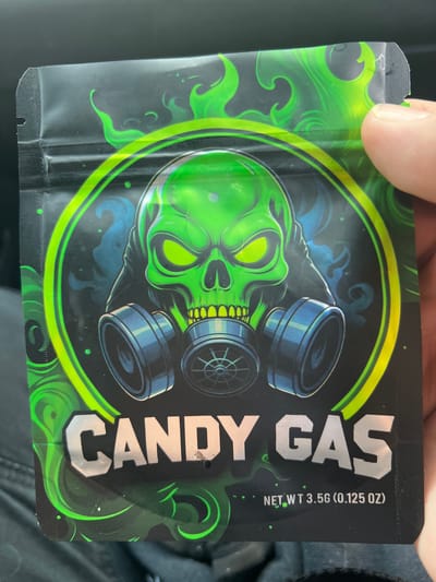 The scene, purportedly located in the Town of New Windsor, United States of America, depicts a close-up, slightly angled view of a person's right hand holding a black, flexible, stand-up pouch. The pouch, oriented vertically, features a prominent graphic design on its front.
The central design element is a highly stylized, luminous green skull depicted wearing a dark blue gas mask. The skull has bright, glowing green eyes and a visible row of yellow-green teeth. The gas mask has two circular filter canisters on the sides, with a central respirator component. This skull and gas mask imagery is encircled by a thick, bright neon-green ring. The background of the design is black, with swirling, flame-like patterns in lighter green and faint, smoky blue elements positioned around the skull.
Below the main graphic, the words "CANDY GAS" are printed in large, reflective silver-colored block letters. Beneath this, in smaller white text, reads "NET WT 3.5G (0.125 OZ)".
The pouch itself appears to be made of a glossy, possibly plastic or foil material. Its top edge features a visible tear notch on the left side and a resealable zipper closure running horizontally, partially obscured by the holding hand on the right. A portion of a second tear notch is visible on the top right edge, where a light-skinned thumb and index finger grip the pouch. The person's hand shows no distinguishing marks beyond visible skin texture and a trimmed fingernail on the thumb.
The background behind the pouch is dark and out of focus, suggesting an interior setting with soft textures, possibly dark fabric from a car seat or similar upholstery, visible primarily in the lower left quadrant of the image.