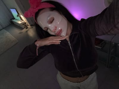 An individual is depicted in an indoor residential setting, consistent with a private residence in Tielrode, Belgium. The subject is a female with long dark hair, wearing a pink bow-shaped headband. Her face is covered by a translucent sheet mask. Her eyes appear closed or mostly closed, and her lips are slightly parted, exhibiting a glossy texture. She is wearing a dark brown, possibly velour, zip-up jacket, partially unzipped to reveal her midriff and a navel piercing. Light-colored pants are visible below her waist. Her right arm is raised, consistent with holding a camera for a selfie. Her left arm is bent, with her hand resting near her left shoulder/chest area, displaying manicured nails and a light pink bracelet on her wrist. The background on the left includes a light-colored rug on a dark floor, and a white console table with a glowing electronic device (tablet or laptop) displaying a green-blue screen, illuminated by a small lamp. A blurry decorative object is beside the lamp. The background on the right is darker, showing a multi-shelved unit. The room is dimly lit with a prominent purple light source emanating from the upper right, casting a magenta hue on the wall.