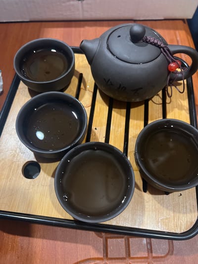 A high-angle shot depicts a tea set arranged on a wooden tray with black edging. The tray rests on a wooden surface. A dark gray teapot with a circular handle is positioned on the left. A decorative knot adorns the handle. Three filled tea cups of the same dark gray color are neatly aligned on the right side of the teapot. One additional tea cup is positioned at the bottom left, completing a set of four cups. A small, black disc is placed beside one of the lower right tea cups. The liquid in each tea cup appears to be brown. The specific location is Ильинская Поляна Снт, Russia.