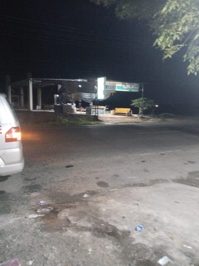 The image depicts a nighttime roadside scene in Meukokuthang, Indonesia. In the foreground on the left, the rear quarter of a light-colored vehicle, likely a minivan, is visible, with its left taillight illuminated. The ground in the immediate foreground consists of a mottled concrete or asphalt surface displaying damp areas and scattered debris, including discarded plastic cups and wrappers. A paved road occupies the midground, showing faint linear markings. Beyond the road, on an elevated terrain, an open-sided building with white support columns and a dark roof structure is partially discernible on the left. Central to the background, a brightly lit sign displays the word "ASIAN" in white capital letters on a dark ground, accompanied by green and orange graphic elements. To the right of the sign, a distinct yellow-orange wooden bench is positioned next to a small tree with some visible foliage. The far background is largely dark, with a faint, distant light source. Overhead, fine horizontal lines, indicative of power lines, cross the dark sky. The upper right corner of the frame contains blurred green foliage from an overhanging tree. The scene is static, with no visible people or discernible activity.