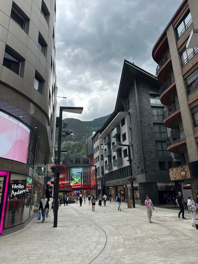 This image captures a bustling pedestrian street in Andorra la Vella, Andorra, under an overcast sky. The street is lined with contemporary multi-story buildings that house various commercial establishments and possibly residential units. In the background, forested mountains are visible, indicating the city's mountainous setting.
Numerous people are seen walking along the paved street, suggesting a lively urban area. Many individuals appear to be shopping or strolling. The general activity is that of a typical city street scene during the daytime, despite the cloudy weather.
Notable architectural details include a building on the left with a distinctive curved, paneled facade and square windows. Buildings on the right feature a mix of dark, textured stone cladding on lower levels and modern window designs, some with curved balconies. Streetlights and hanging baskets with flowers are also visible, adding to the urban environment.
Visible text and brands on signage and digital displays include: "Hello Andorra," "Elizabeth Arden," "NOUS COFRES DE REGAL," "OYSHO," "UAILO'S Food," "UAILODUTY FREE," "Intimissimi," "KIKO," and a partially visible "COMPANIA ANDORRANA D'ASSE." The cloudy sky suggests a cool or mild day. The scene evokes a sense of modern urban life amidst a natural, mountainous landscape.