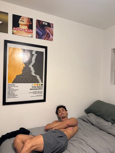 A male subject is depicted lying on his back on a bed in a room located in Bondi, Australia. The subject, an adult male with dark, curly hair, a mustache, and stubble, is shirtless and wearing dark grey athletic shorts. His forearms are crossed over his torso as he looks directly towards the camera with a neutral expression. The bed is covered with grey bedding, and a grey pillow is positioned against the white wall on the right. A crumpled black garment lies on the bed to the subject's left. The white wall behind the bed is adorned with several items. Three album covers are mounted above the bed: a partially visible gold cover on the far left, "Radiohead - The Bends" in the center, and "RÜFÜS DU SOL" on the right. Below these, a large framed poster for "Tom Misch - Geography" displays an orange and dark grey silhouette design with a tracklist and release information. A small window with a white frame and white blinds is visible on the upper right wall.