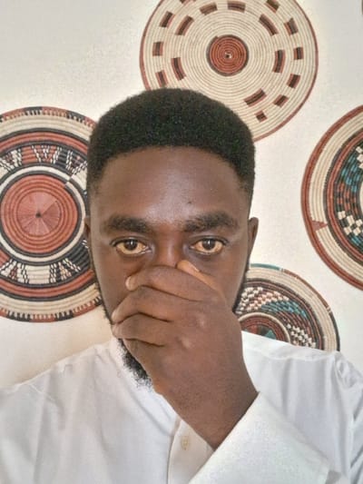 An adult male of African descent is depicted from the mid-chest upwards, situated indoors in Takum, Nigeria. He possesses short, dark, textured hair and a sparse beard. His visible eye sclerae exhibit a distinct yellow discoloration. He wears a white, collared, long-sleeved shirt. His right hand is placed over his mouth and nose, with fingers slightly curled. The background wall is light-colored and adorned with three circular, woven decorative wall hangings. The top-left and top-right hangings feature concentric patterns in various shades of brown and beige. A third partial hanging is visible on the lower right, displaying patterns that include blue and brown tones.