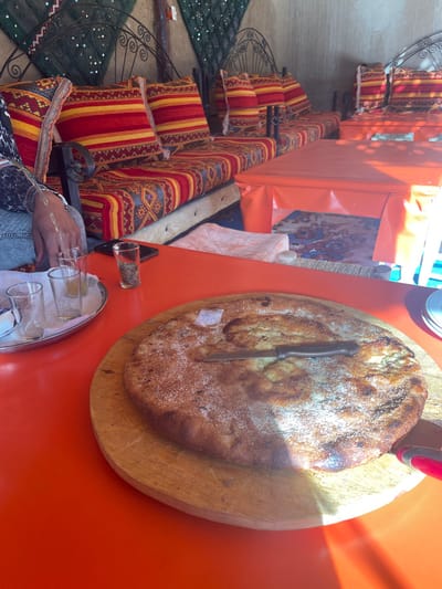 An overhead shot shows a large, circular flatbread dusted with what appears to be sugar, stabbed with a knife on a wooden board. An orange table holds the bread and a tray with several small glasses. A person's arm and hand appear to pour a liquid into one of the glasses. Cushioned seating with patterned fabric lines the side of the table, suggesting an outdoor cafe or market setting. The scene is brightly lit, indicating a sunny day.