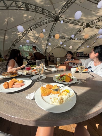 The image depicts a group of people enjoying a meal at a table, seemingly outdoors under a large, tent-like structure with a geodesic dome roof supported by metal beams. Several round, white and orange lanterns are suspended from the ceiling. Sunlight filters through the translucent roof, casting shadows on the table and the floor.

The focus is on a table laden with food and drinks. Plates hold various breakfast items, including fried eggs, hash browns, sausages, sandwiches, and a salad with cheese. Wine glasses, some filled with white wine and one with a red liquid, are also present. Two women are visible in the foreground, one with dark hair looking towards the table, and another with long dark hair reaching for a glass. A young child wearing a baseball cap is seated between them, interacting with something on the table. Other people are blurred in the background, suggesting a more general gathering or restaurant setting.

The scene appears to be during the day, with bright sunlight indicating fair weather. The presence of a tent structure and the natural lighting suggest an outdoor or semi-outdoor dining experience. The overall atmosphere seems relaxed and casual, with people engaged in eating and socializing. The location context is provided as Morozovo, Russia. There is no visible text in the image.