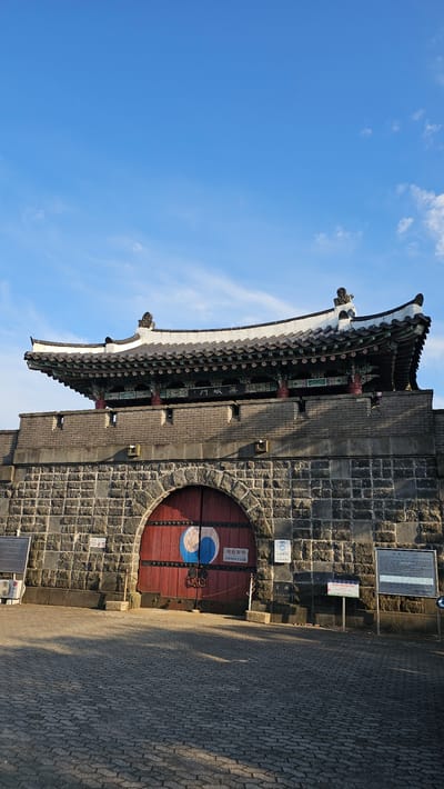 The image shows a wide-angle view of a large, stone gate with a traditional Korean roof. The gate is made of rough-hewn, dark grey stones and features a large, arched, red wooden door. On the door, there is a circular symbol in blue and white. Above the gate, a multi-tiered, dark roof with white trim and decorative eaves is visible. The roof is supported by ornate, painted pillars. The gate is part of a larger stone wall, which is also constructed from similar grey stones. In front of the gate, there is a paved area made of interlocking bricks, which casts long shadows from an unseen light source, suggesting it is either morning or late afternoon. Several signs are posted on the wall to the right of the gate, some with Korean text and one with a blue and white logo. The sky above is a clear, bright blue with a few wispy white clouds. This is likely a historic site, possibly a fortress gate, located within the city of Cheonan-si, South Korea.