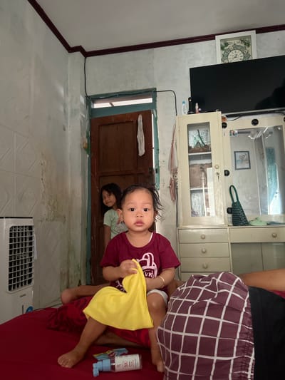 The image depicts an indoor scene, likely a bedroom, in Kecamatan Pati, Indonesia. In the foreground, a young child, possibly a toddler, sits facing the camera, holding a yellow cloth. The child is wearing a maroon t-shirt and white shorts. To the left of the child, a white portable air cooler is visible. Behind the child, a slightly older girl stands peeking out from behind a wooden doorframe, wearing a light green t-shirt. The room's walls are a pale color, with some water damage and what appears to be textured wallpaper on the left side. A large television is mounted on the wall above a white cabinet with glass doors and drawers. A framed clock is visible on the wall above the television. The floor is covered with a red surface, likely a bed or mattress, on which the children are seated. Various objects, including a bottle of hand sanitizer, are scattered on the red surface. The overall impression is of a casual, domestic environment.