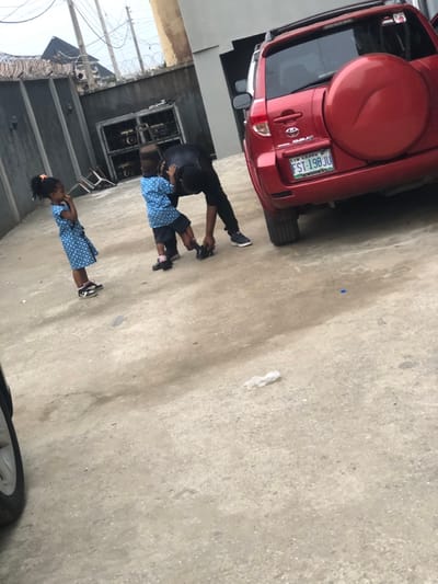 The image is rotated approximately 90 degrees clockwise, depicting an outdoor scene with an adult and two children on a paved surface, alongside a parked red SUV.

In the foreground, an adult, dressed in a dark shirt and dark trousers, is leaning down, appearing to assist a child with their footwear. The child, likely a boy, is positioned directly in front of the adult, facing away from the camera. This child wears a blue and white patterned uniform, consisting of a short-sleeved top and shorts, paired with dark sneakers.

A second child, likely a girl with dark hair tied back, stands a short distance to the right of the adult and first child. She is also dressed in an identical blue and white patterned uniform, including a dress or top and skirt, and dark sneakers with light colored soles and pink accents. She is facing towards the left side of the frame, observing the interaction.

To the left side of the frame, a red Toyota RAV4 SUV is parked. The vehicle has a spare tire mounted on its rear door. A visible license plate reads "FST-198 JU," with "OYO - STATE" printed beneath the numbers.

The ground is a light-colored concrete or paved surface, showing some minor stains or dirt. A small blue cap and a crumpled white piece of paper or plastic are visible on the ground.

In the background, on the far left, there is a grey building or wall, and a fence topped with barbed wire is partially visible. Further back, utility poles and wires are present against a light sky. Behind the children, another building or structure features dark, rectangular openings that resemble windows or a gate.

The scene appears to be taking place during the daytime, under natural light, suggesting a casual moment of an adult helping children, possibly getting ready for school or an outing given their uniforms.