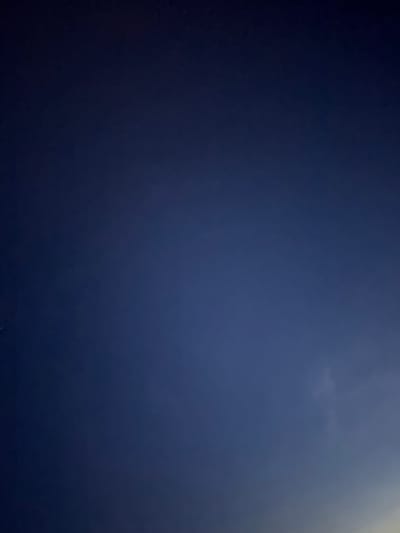 This is a photograph of a dark blue sky with a gradient of lighter blue and some faint white cloud formations visible in the upper right quadrant. There are a few very small, faint white dots, possibly stars or distant lights, scattered across the frame, particularly in the lower and mid-left areas. The overall impression is of a twilight or night sky. The lighting suggests a source of light from the upper right, which is creating a slight luminous effect on the clouds and the sky. There is no discernible text, people, or specific objects in the image, and no active event is taking place. The scene is entirely focused on the sky itself. Given the location context, this could be a view of the sky above Володарский ауылдық округі, Kazakhstan, likely during the evening or early morning hours when the sky is not fully dark.