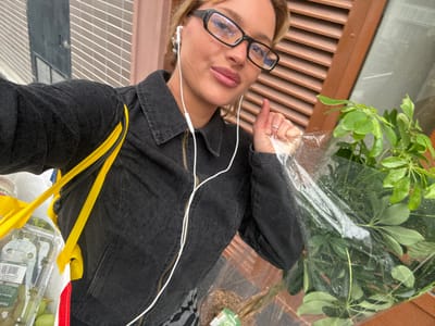 A young adult female is depicted in a close-up selfie on a street in Nogent-sur-Marne, France. She has light brown hair, wears black-framed glasses with blue anti-glare coating, and white wired earphones. She is dressed in a dark, possibly black, denim jacket with a zippered front. Her right hand, adorned with a ring on the ring finger, holds a green potted plant encased in clear plastic. A yellow fabric bag hangs over her left shoulder, containing a clear plastic container of green grapes. The background on the left features a building with light-colored horizontal siding and a dark window, while the right side shows a building with reddish-brown horizontal slatted shutters.