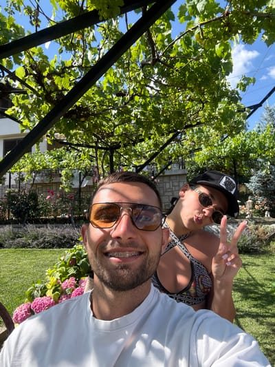 A man and a woman are taking a selfie outdoors in a garden setting. The man, in the foreground, is wearing a white t-shirt and sunglasses. He has braces on his teeth. The woman, behind him, is wearing a patterned bikini top and a baseball cap. She is making a peace sign with her hand and puckering her lips. The background shows a lush garden with green foliage, pink flowers, and a vine-covered trellis. The scene appears to be in a residential area, possibly in the city of Chavei, Bulgaria, given the architecture and landscaping. The sky is blue with some clouds.