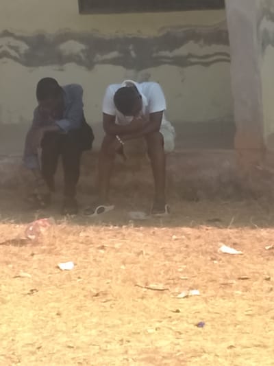 The image depicts two young males seated on a concrete ledge against a discolored, light-colored wall in Nassarawa Egon, Nigeria. The male on the left wears a dark blue long-sleeved shirt and dark trousers, with his head bowed and resting on his left arm, supported by his left knee. The male on the right wears a white short-sleeved polo shirt and light-colored shorts, with his head bowed, hands clasped, and resting on his knees. A white beaded bracelet is visible on his right wrist. White slide sandals are placed on the dry, sparse, light-brown vegetation covering the ground in front of him. Scattered litter, including a red plastic bag and white paper fragments, is visible on the ground. The wall behind the subjects shows dark, wavy discoloration patterns.